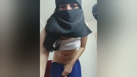 Video of a slender, light-skinned woman with long black hair, partially obscured by a black veil, wearing a white crop top and blue pants.