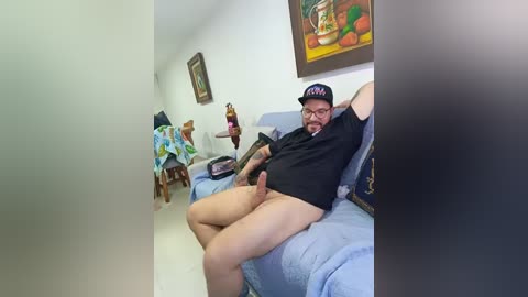 A video of a bearded man with a large, erect penis, wearing glasses, a cap, and a black shirt, lying on a blue sofa in a dimly-lit room with colorful artwork on the wall.