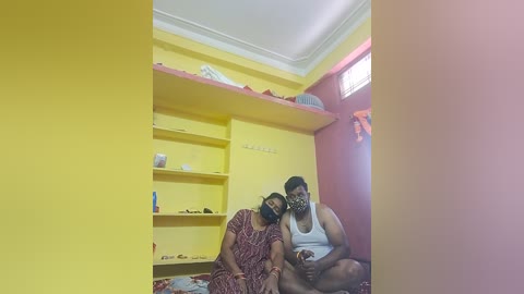 Video of two people in a small, brightly lit room with yellow walls. A woman in a red dress and a man in a white tank top sit on the floor, both wearing face masks. Shelves hold items.