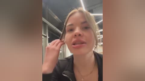 Video of a young, fair-skinned woman with blonde hair, wearing a black leather jacket, adjusting her hair in a modern, industrial setting with exposed pipes and bright lights.