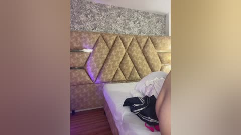 Video of a modern hotel room with a tufted gold headboard, floral-patterned wallpaper, and a neatly made bed with white linens and a black jacket on the right.