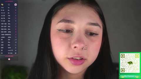 A close-up video of a young woman with fair skin, long black hair, and a silver nose ring, looking introspective. A digital calendar and a QR code are visible in the background.