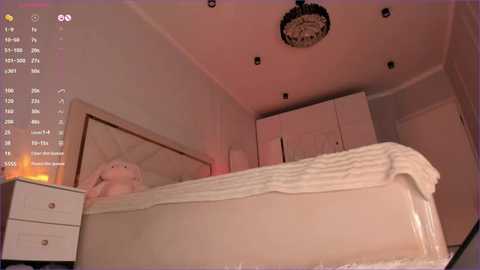 A video of a dimly lit bedroom with a large bed, white bedding, and a fluffy white pillow. A chandelier hangs from the ceiling, and a digital clock display is visible in the upper left corner.