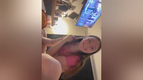 Media: Video of a smiling young Asian woman with long black hair, wearing a pink top, sitting at a cluttered desk with a computer monitor displaying a game screen.