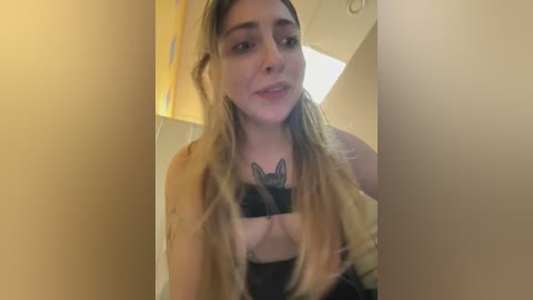 A video of a smiling young woman with long blonde hair, wearing a black bra with a bat emblem, standing in a dimly lit room with beige walls and a yellow lamp.