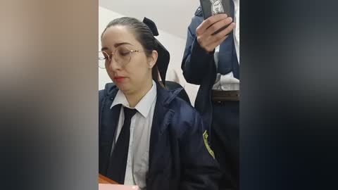 Video of a young woman with glasses and a black headband, wearing a navy jacket over a white shirt and black tie, seated at a table, possibly in a courtroom.
