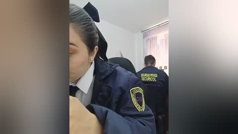 Video of a woman with dark hair tied back, wearing a blue school uniform jacket, seated in a classroom; two uniformed school security guards in the background.