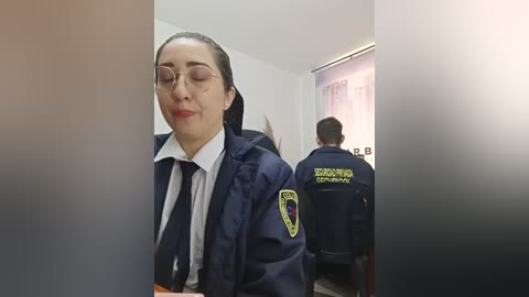 Video of a female police officer with glasses and a navy jacket, standing in a hallway with another officer behind her.