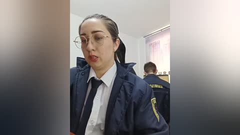 Video of a woman in a navy-blue police jacket, wearing a white shirt and glasses, standing in a dimly lit room with two other officers in the background.