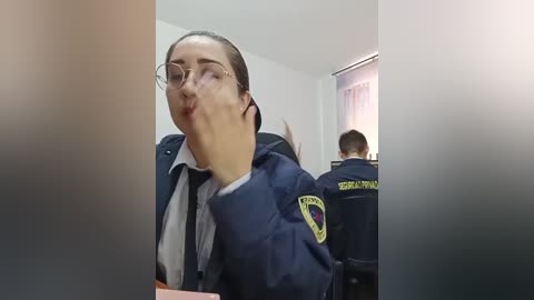 Video of a woman with glasses and dark hair, making a goofy face, standing next to a uniformed security guard in a brightly lit room.