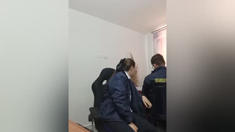 Video of two police officers in dark blue uniforms, one with \"BRI\" marked on the back, questioning a man seated in a black gaming chair, in a sparse, white-walled room with a window.