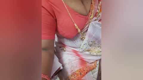 Media: A video of a woman wearing a vibrant, red short-sleeved top and a colorful, floral saree, adorned with a gold necklace and bracelets. The image is slightly blurred, focusing on her torso and upper arms.