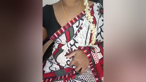 Video of a South Asian woman with medium brown skin, wearing a black blouse and a vibrant, patterned sari with red, white, and black floral designs. She holds a white garland with red beads in her right hand.