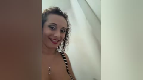 Video of a smiling, curly-haired woman with medium breasts in a black-and-white striped top, standing in a dimly-lit hallway.