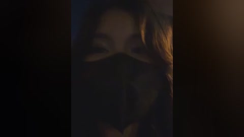 A dimly lit video featuring a partially obscured woman with long, wavy hair, wearing a dark mask, against a dark background, creating an eerie, mysterious atmosphere.