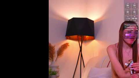 A video showing a young woman with long brown hair wearing a pink top, holding a glass, in front of a black lamp with a warm glow, in a cozy indoor setting.