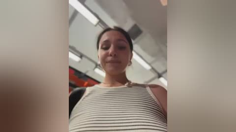 Video of a young Asian woman with dark hair in a ponytail, wearing a white and black striped tank top, smiling, indoors with fluorescent lighting and blurred background.