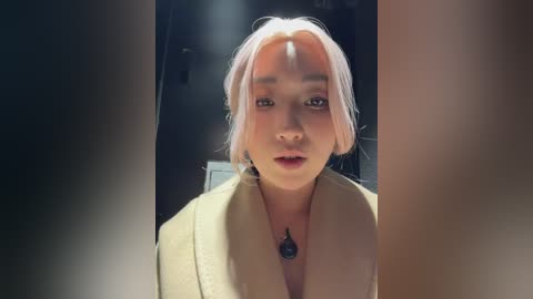 Video of a young East Asian woman with fair skin and pastel pink hair, wearing a beige coat, standing in a dimly lit room with blurred background.