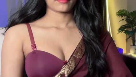 Video of a South Asian woman with medium skin tone, wearing a maroon bra with a gold shoulder strap, long black wavy hair, smiling with red lipstick, indoor background with a potted plant and neutral decor.