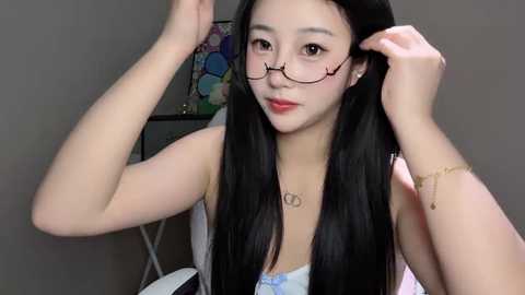 Video of an East Asian woman with long black hair, wearing glasses, adjusting them while seated indoors. She has a fair complexion and is dressed in a light blue top.