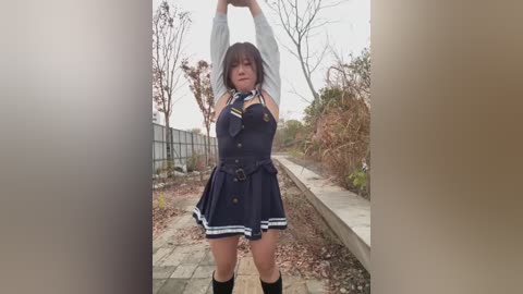 Video of a young woman with medium-length brown hair, wearing a navy blue dress with white stripes, standing outdoors on a stone path, stretching her arms above her head. Background includes bare trees and a metal fence.