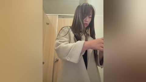 A video of a young Asian woman with long, straight black hair, wearing a white robe, standing in a bathroom with beige walls and a wooden door.