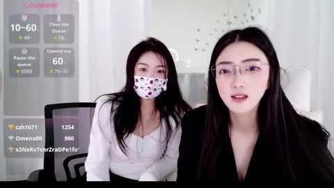 Video of two young Asian women, one masked, in a modern office setting with white walls and a digital screen displaying streaming statistics.