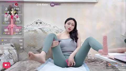 Video of an Asian woman with long black hair, wearing a gray tank top and tight green leggings, sitting on a bed, legs spread, with a large pink dildo, in a bedroom with a white headboard.