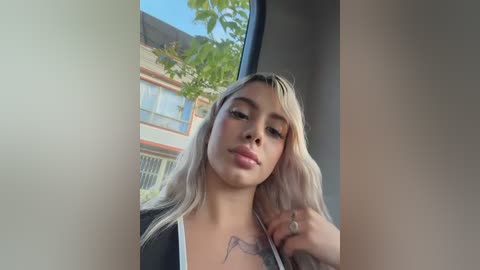Video of a blonde woman with long, wavy hair, light skin, and a tattoo on her chest, posing inside a car with a modern building and trees visible through the window.