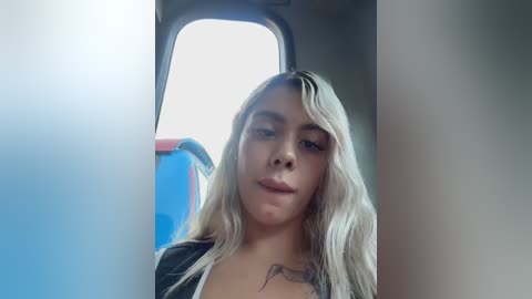Video of a blonde woman with light skin, wearing a black top, seated in a bus. She has a tattoo on her chest and looks towards the camera. Background shows bus seats and window.