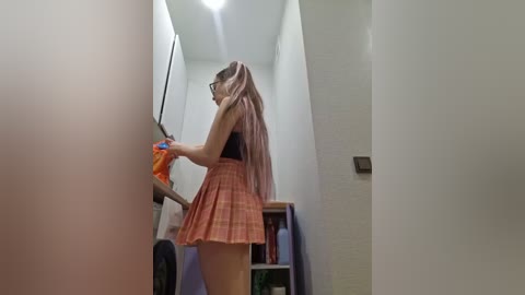 Video of a young woman with long pink hair in a plaid skirt and black top, washing dishes in a narrow kitchen with white walls and cabinets.