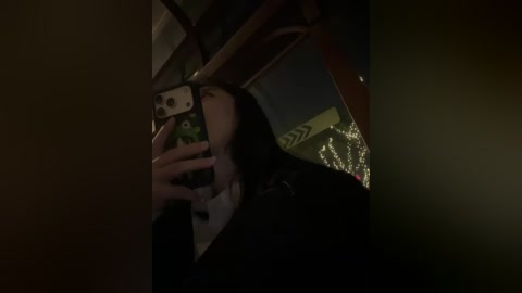 A dimly lit video captures a young woman with long dark hair taking a selfie in a car. She holds a phone with a green case, partially visible in her hands. The background reveals a dark, cluttered interior.