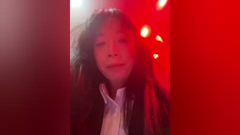 Video of a young Asian woman with long black hair, wearing a white shirt, against a red-lit background, smiling slightly, with a soft focus.