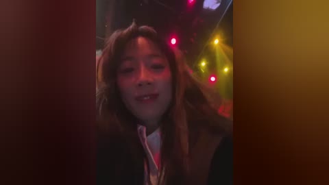 A video of a young, smiling African-American woman with shoulder-length, wavy hair, wearing a dark coat, standing in a dimly lit, festive environment with colorful, glowing lights in the background.