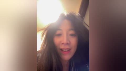 Video of a woman with long brown hair, smiling, in a dimly lit room with warm lighting. She is slightly out of focus, with a soft glow around her head.