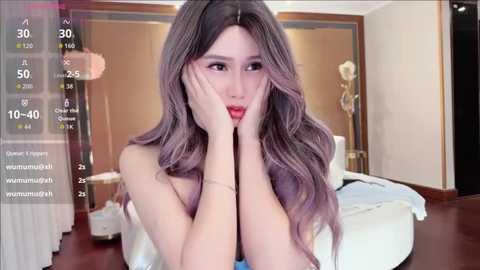 A video of a young East Asian woman with long, wavy purple hair, wearing a white top, sitting in a modern bedroom with a bed, wooden floor, and beige walls.