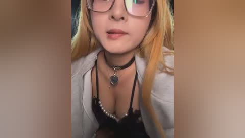 Video of a fair-skinned, blonde woman with glasses, wearing a black lace bra, black choker, and open light jacket. Background is blurred, focusing on her cleavage.