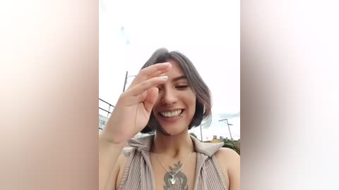 Video of a young woman with short dark hair, wearing a striped top, smiling, and shielding her eyes with her hand, set outdoors against a cloudy sky.