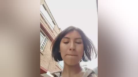 Video of a young woman with straight, shoulder-length black hair and closed eyes, standing in front of a brick building with windows. The image is slightly blurry.