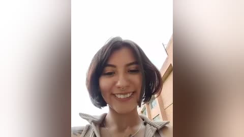 Video of a young woman with short, dark hair, light skin, and a nose ring, smiling, set against a blurry urban background with beige walls.