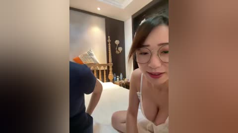 Video of a young Asian woman with fair skin, large glasses, and a white bra, kneeling on a bed in a dimly-lit, cozy room with a wooden headboard.