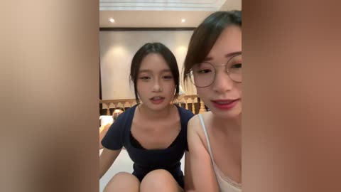 Video of two Asian women with long dark hair, one in glasses, sitting on a bed in a modern bedroom, smiling.