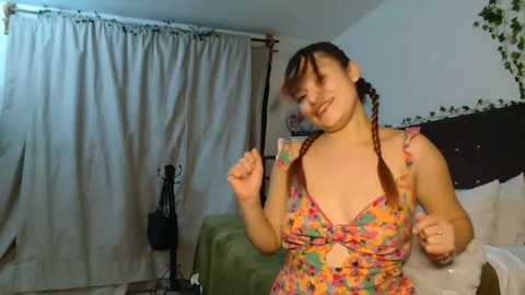 Video of a smiling, light-skinned woman with braided red hair, wearing a colorful floral dress, standing in a simple bedroom with beige curtains and a green bedspread.