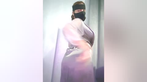 Video of a person with pale skin, wearing a black face mask and a white lab coat, standing in a dimly lit, clinical setting.