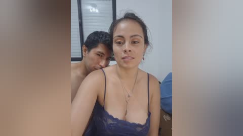 Video of a young, Latina woman with medium-brown skin and dark hair in a side ponytail, wearing a revealing, navy blue lace bra, sitting in a room with a man behind her.