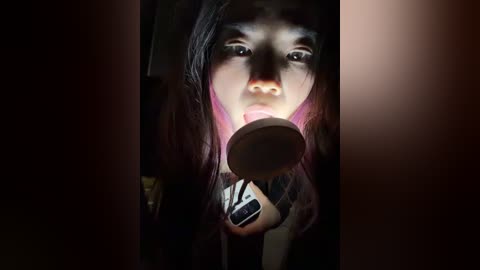 A video of a young woman with long, dark hair, wearing a striped shirt, intensely focused while holding a lit candle close to her face, casting dramatic shadows.
