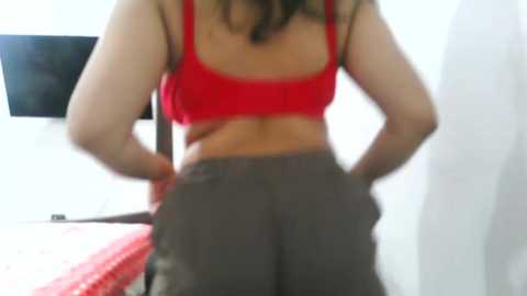 Media: Video of a woman's back in a red sports bra and loose gray sweatpants, standing in a dimly lit room with a TV on the wall and a red blanket on a bed.