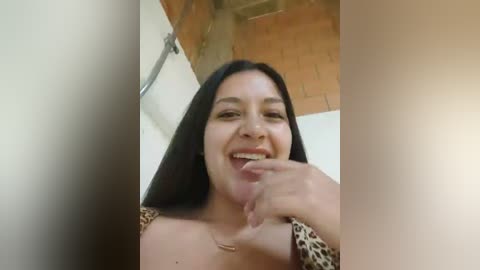 Video of a smiling Latina woman with long black hair, medium skin tone, wearing a leopard-print top, biting her finger, in a rustic room with brick and white walls.