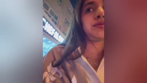 A video of a young woman with long brown hair, partially obscured by a pink wall, wearing a beige jacket with a white floral pattern. The background features a blurred, lit-up sign reading \"WELCOME.\