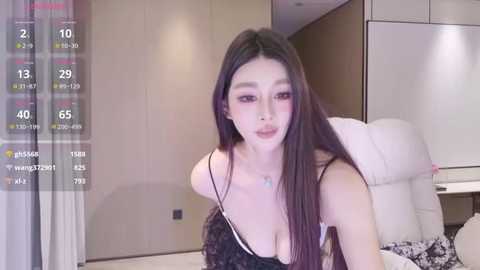 A video of an East Asian woman with long, straight black hair, wearing a black lace dress, in a modern, minimalist bedroom.